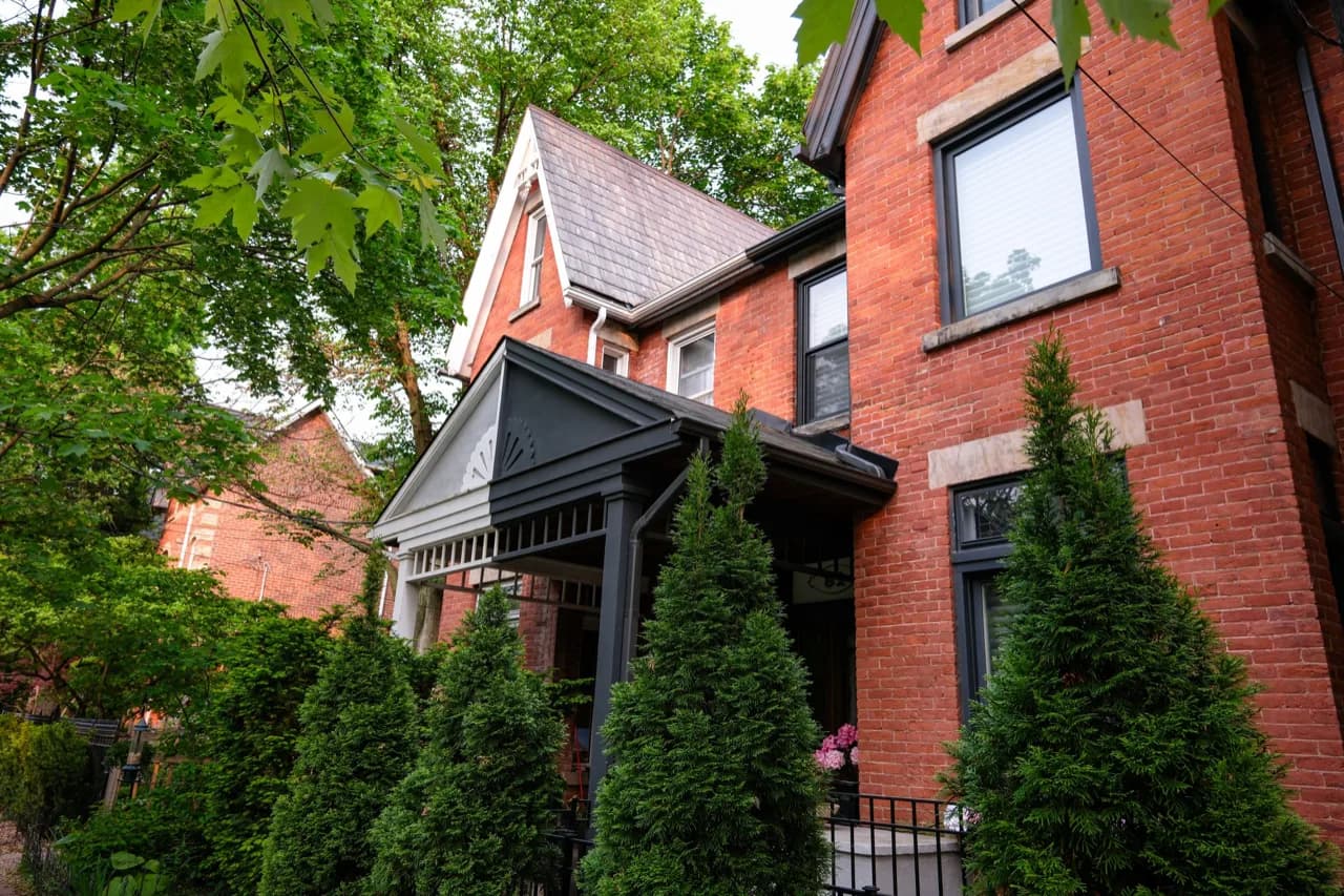 A leafy East York street with a red-brick Victorian house behind cedar shrubs - ADU builders serving East York, Ontario, Canada